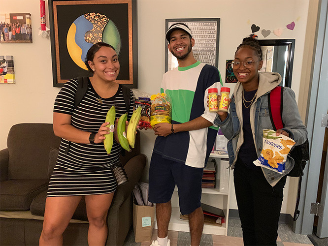 Three students holding donated items, including plantains and various snacks.
